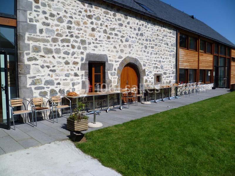 Terrasse avec tables et chaises alignées devant un bâtiment en pierre et bois, bordée par une pelouse verte.