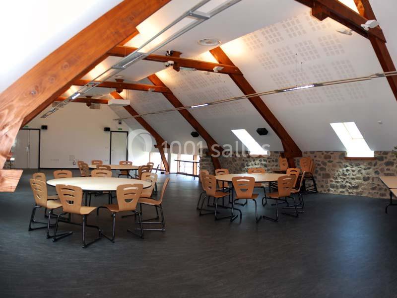 Salle lumineuse sous combles avec poutres apparentes, tables rondes et chaises disposées pour une réunion ou un événement.