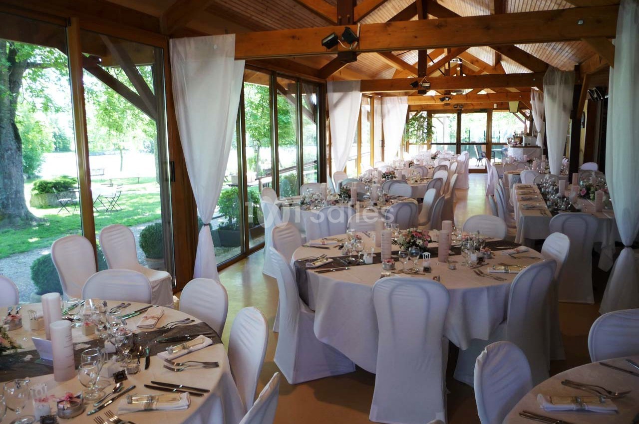 Salle de réception lumineuse avec tables rondes dressées, nappes blanches et grandes baies vitrées donnant sur un jardin.