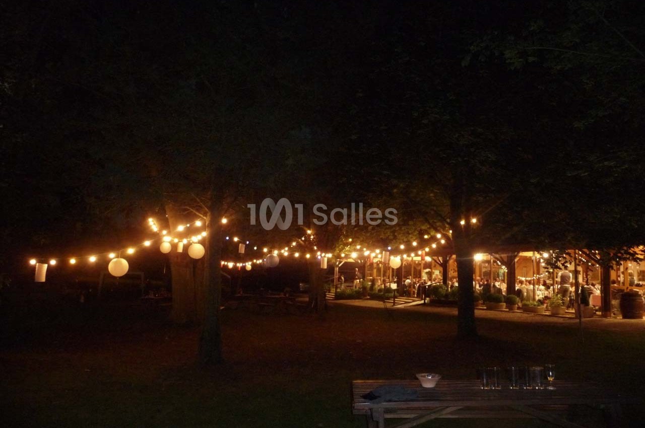Guirlandes lumineuses suspendues dans un jardin sombre, éclairant des tables et une structure en arrière-plan.