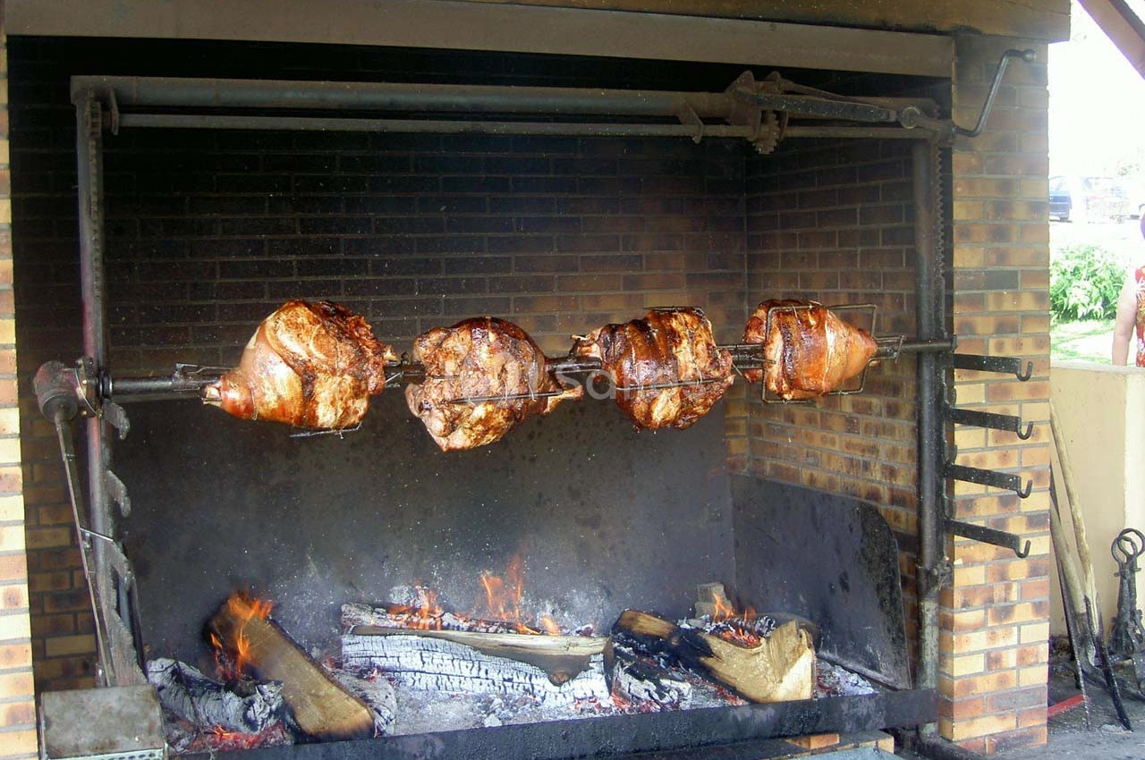 Quatre morceaux de viande rôtissent sur une broche au-dessus d'un feu de bois dans un foyer en briques.