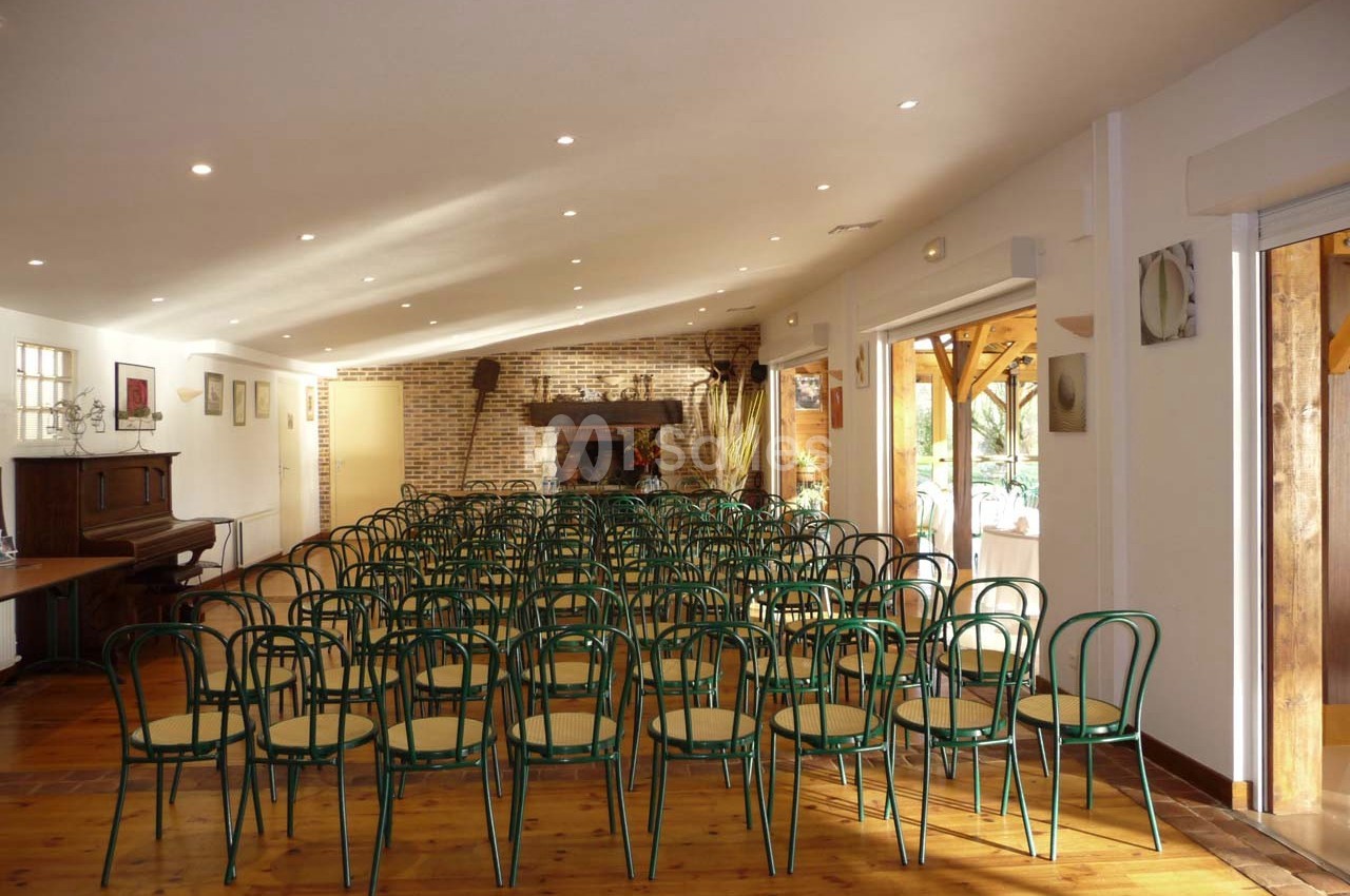 Salle lumineuse avec des rangées de chaises vertes en bois, un piano à gauche et une cheminée en briques au fond.