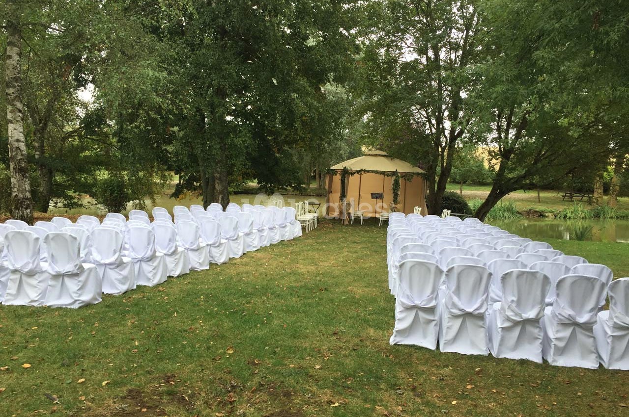 Chaises blanches disposées en deux rangées sur une pelouse, face à une tente beige entourée d'arbres.