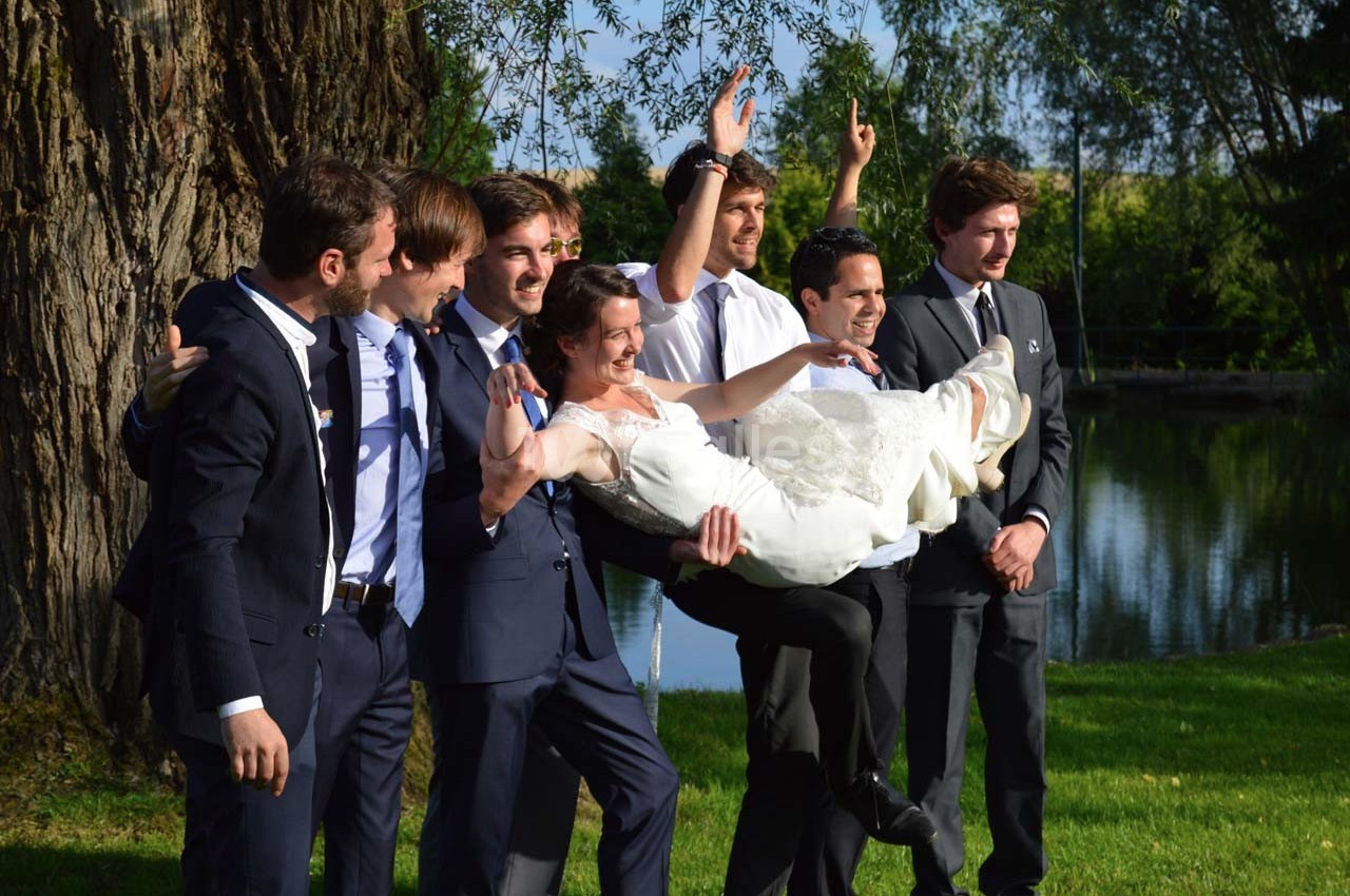 Un groupe d'hommes en costume porte une femme en robe de mariée dans un parc verdoyant près d'un plan d'eau.