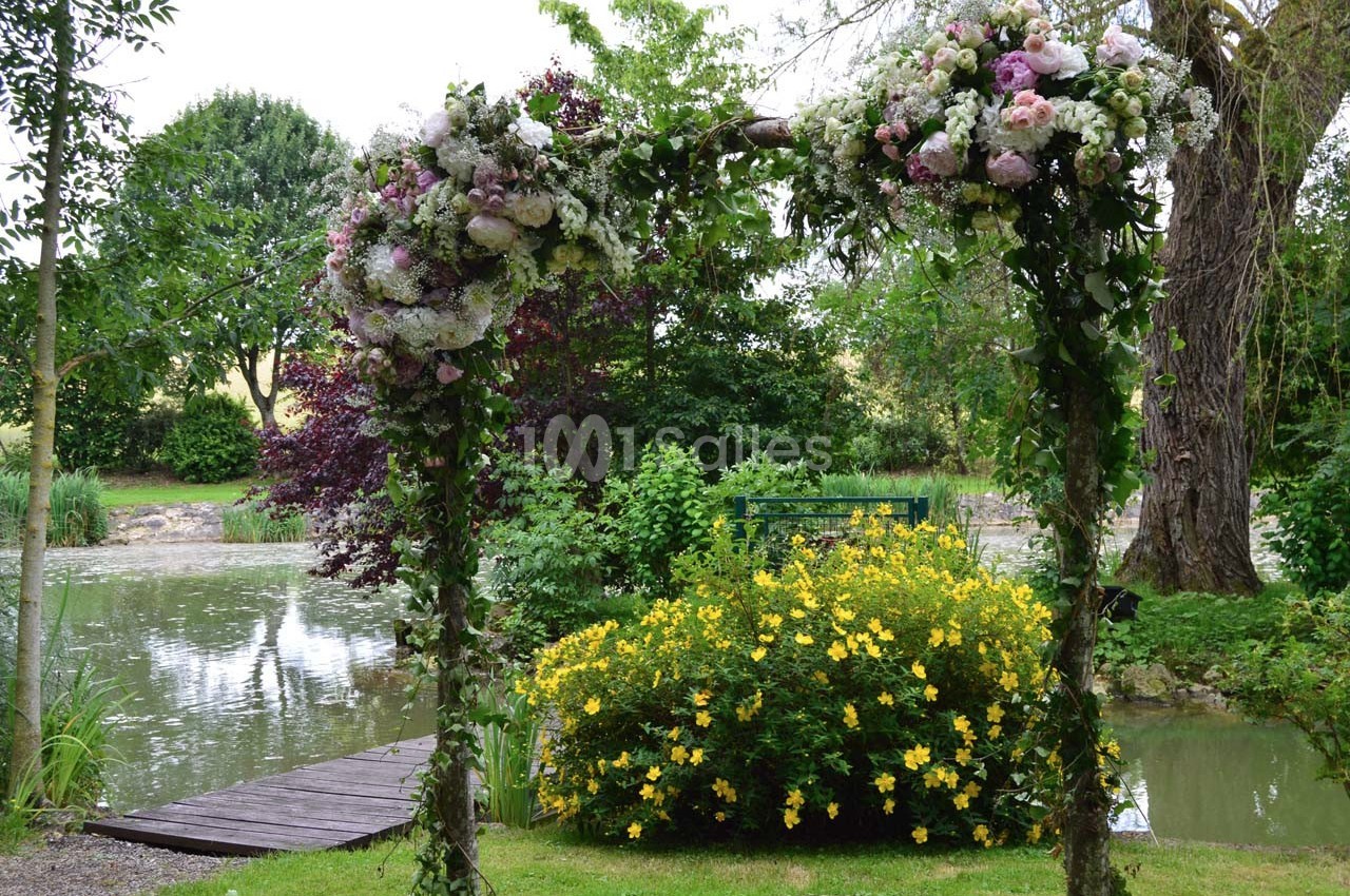 Arche fleurie décorée de roses et feuillages, placée près d'un étang entouré de végétation et de fleurs jaunes.