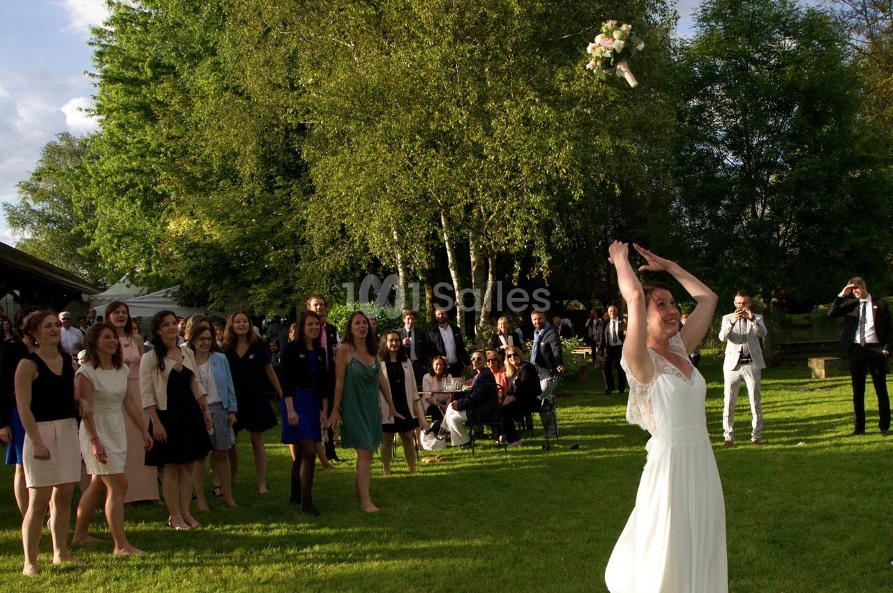 Une mariée lance son bouquet de fleurs à un groupe d'invités rassemblés dans un jardin verdoyant.