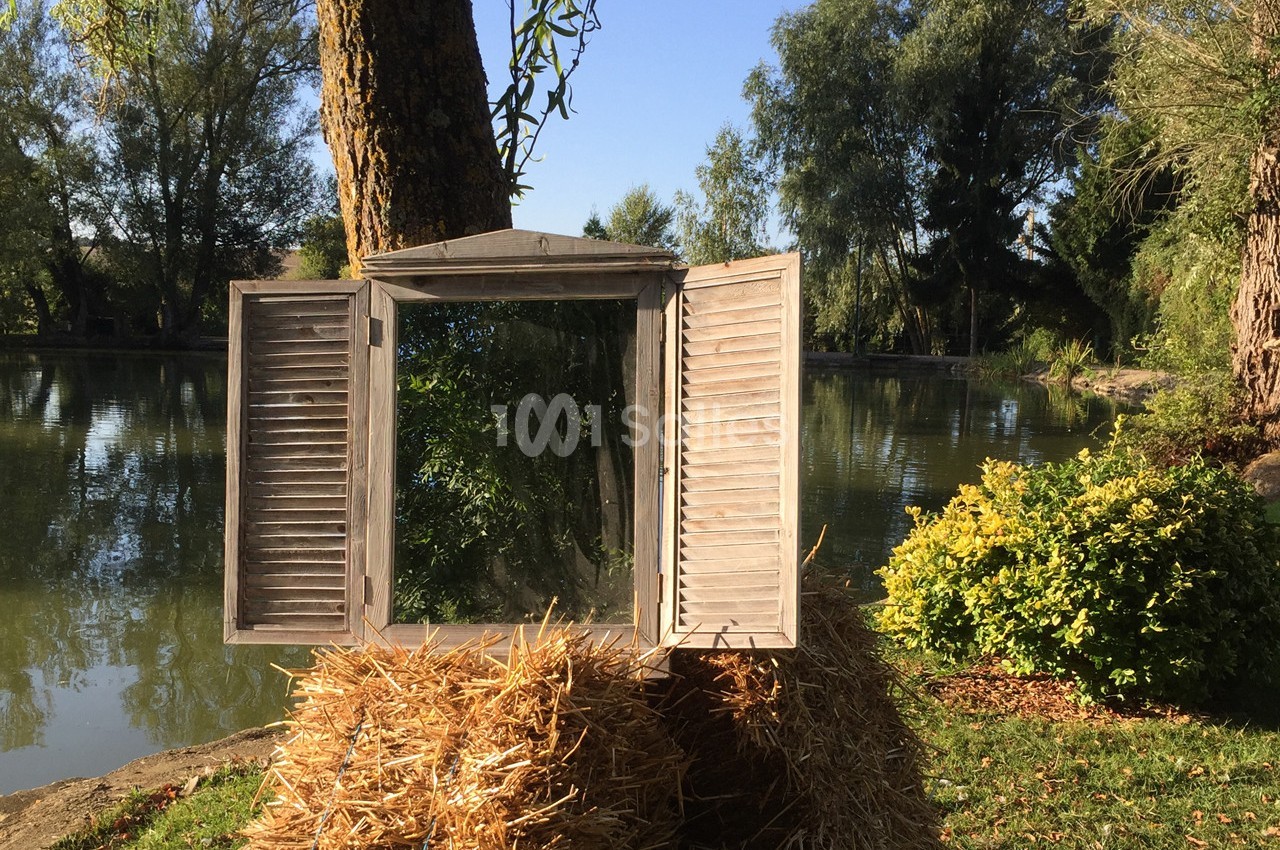 Miroir encadré de volets en bois, posé sur une botte de paille près d'un étang entouré de végétation.