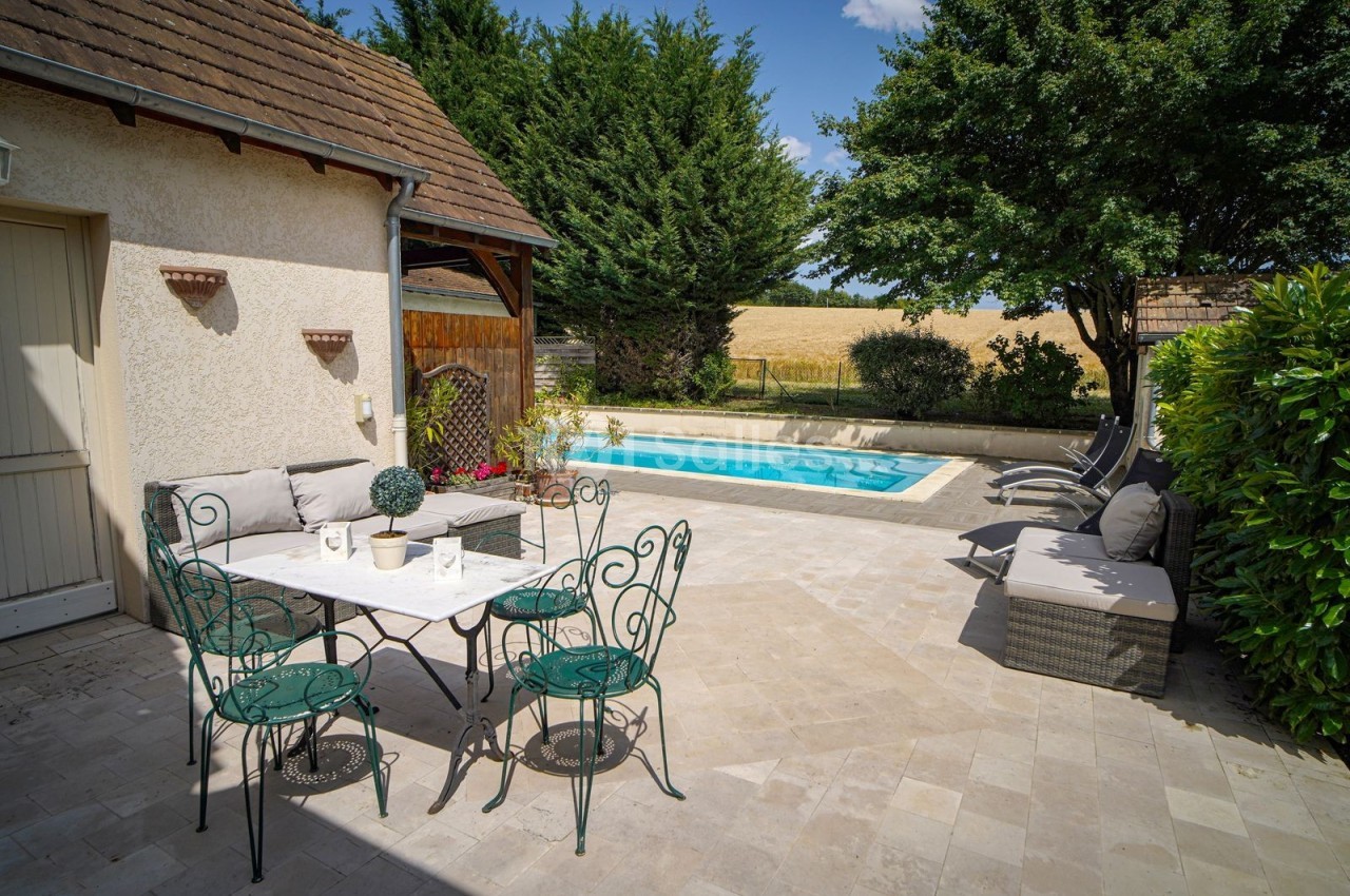 Terrasse avec table et chaises en fer forgé, piscine entourée de transats, arbres et champs en arrière-plan.