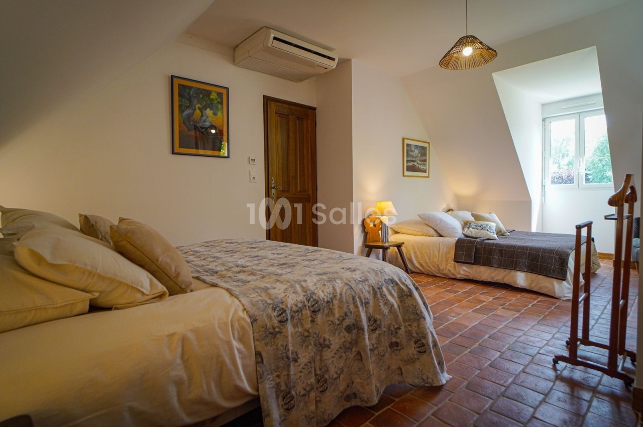 Deux lits dans une chambre lumineuse avec sol en terre cuite, murs blancs, tableaux et fenêtre donnant sur l'extérieur.