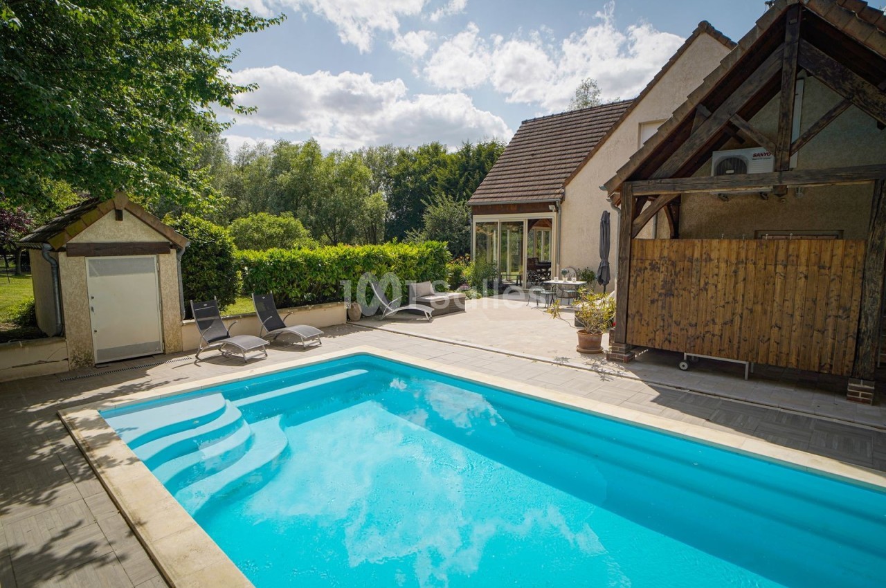 Piscine extérieure entourée de chaises longues, terrasse en bois et maison avec jardin arboré en arrière-plan.