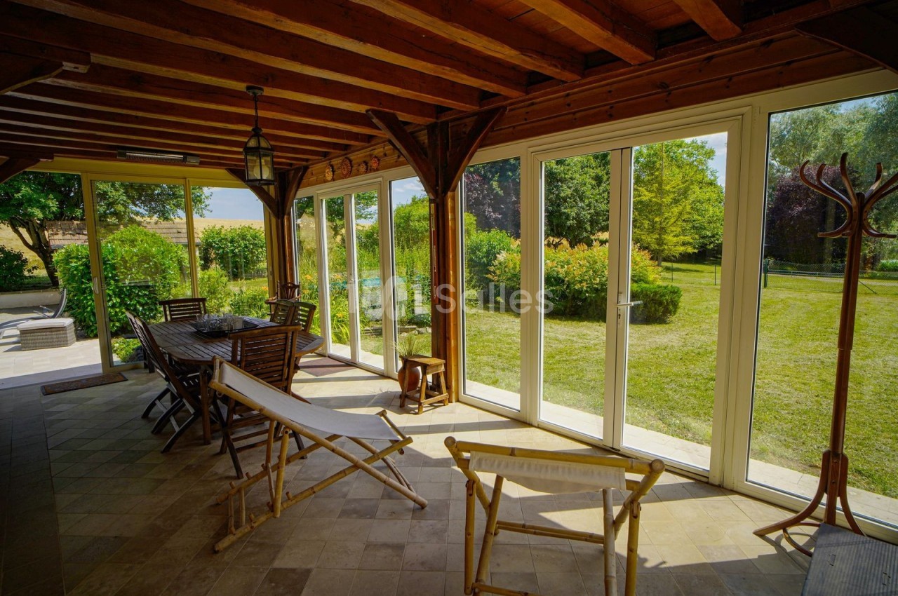 Véranda lumineuse avec mobilier en bois, donnant sur un jardin verdoyant par de grandes baies vitrées.