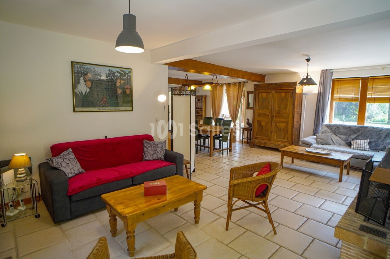 Salon lumineux avec canapé rouge, fauteuils en osier, table basse en bois et vue sur une salle à manger en arrière-plan.