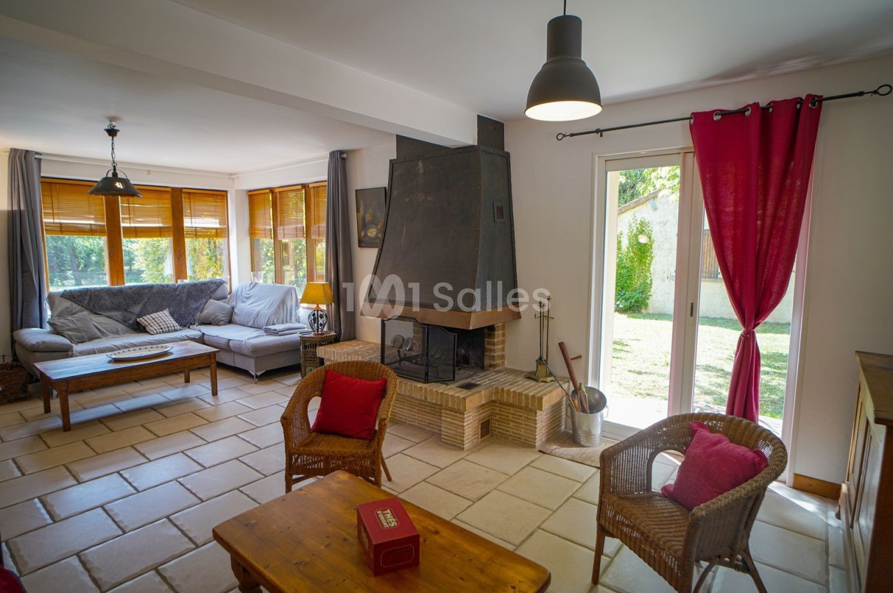 Salon lumineux avec cheminée, canapé d'angle, fauteuils en osier, table basse en bois et vue sur un jardin verdoyant.