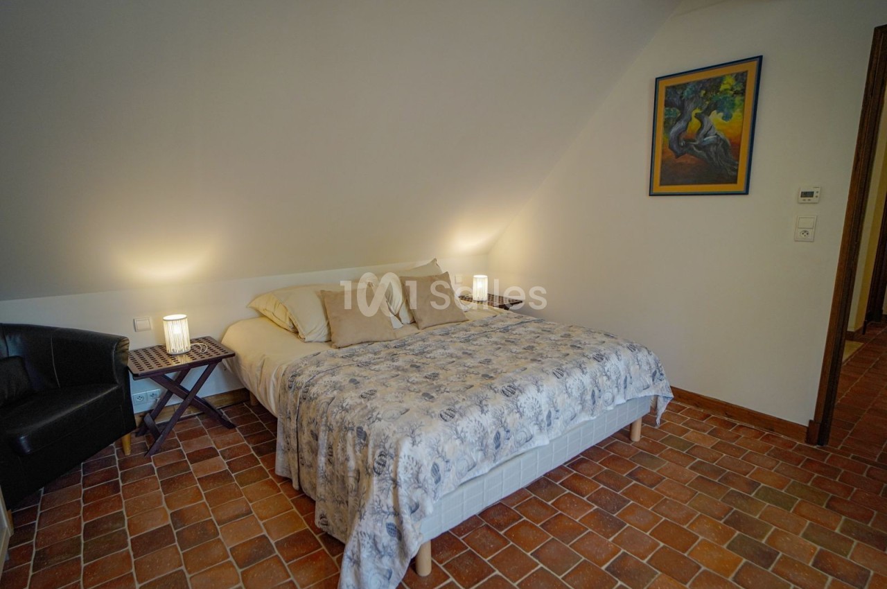 Chambre avec lit double, couvre-lit fleuri, sol en terre cuite, fauteuil noir et éclairage doux.