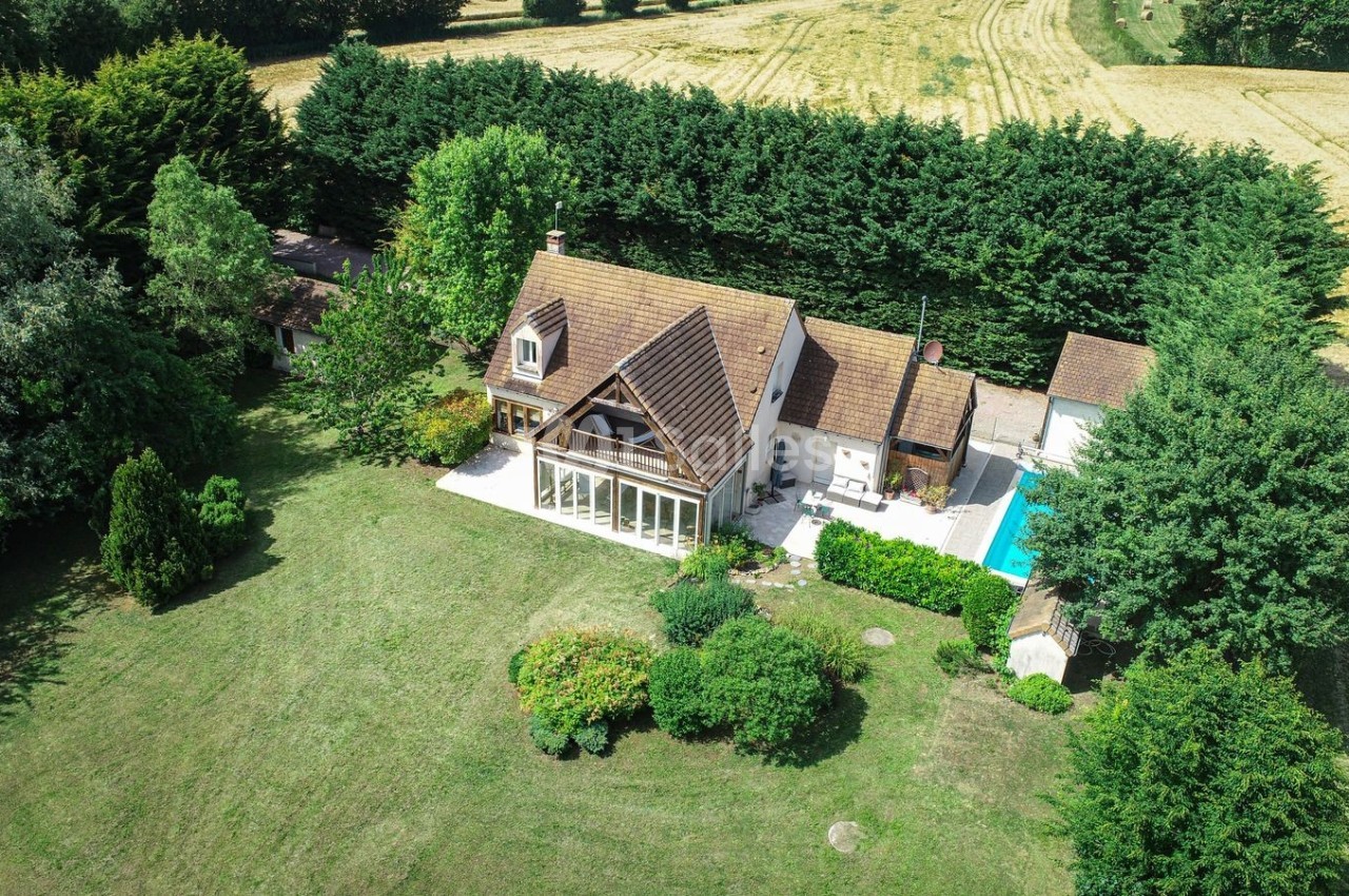 Vue aérienne d'une maison avec toit en pente, jardin arboré, piscine et champs environnants.