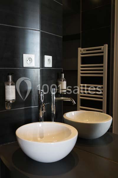 Deux lavabos blancs ronds sur un plan noir, avec un robinet chromé, un miroir et un sèche-serviettes mural.