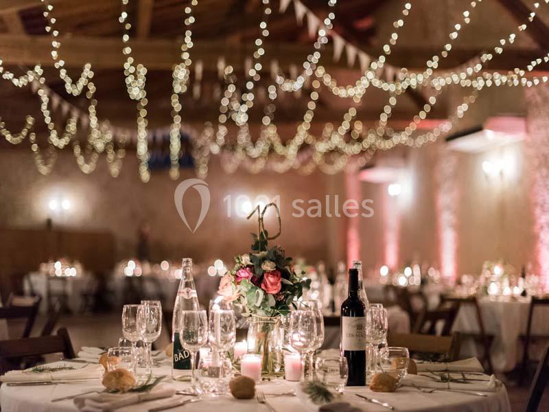 Salle de réception décorée avec des guirlandes lumineuses, tables dressées avec fleurs, bougies et bouteilles de vin.