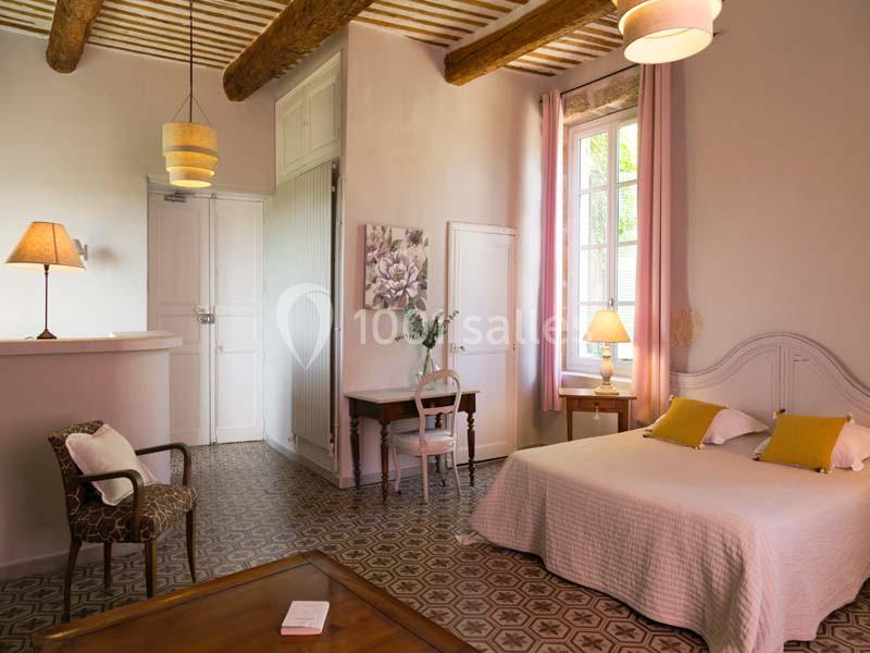 Chambre lumineuse avec lit double, murs clairs, poutres apparentes, mobilier en bois et grande fenêtre avec rideaux roses.