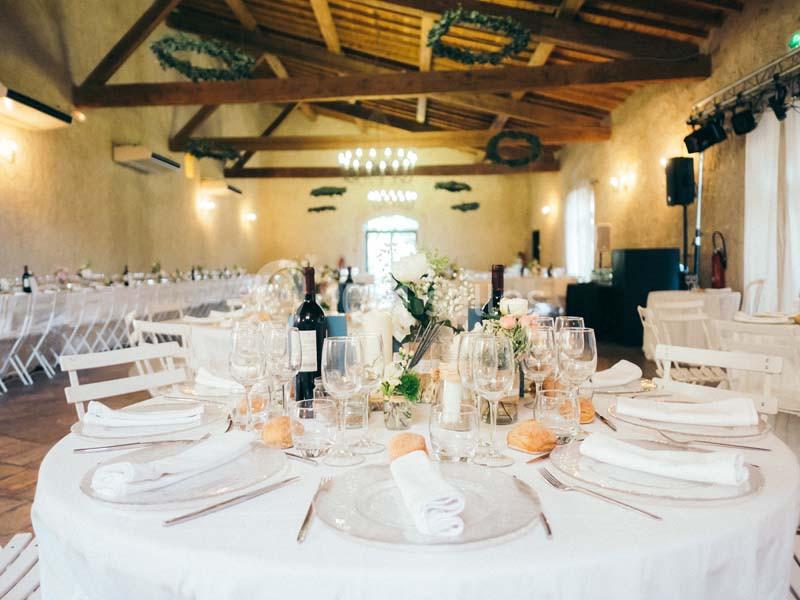 Salle de réception lumineuse avec tables décorées de nappes blanches, vaisselle élégante et fleurs.