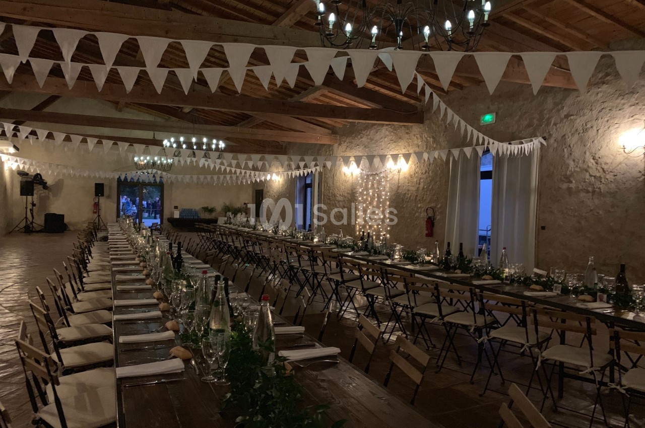 Grande salle décorée pour un événement, avec longues tables en bois, guirlandes lumineuses et fanions suspendus.