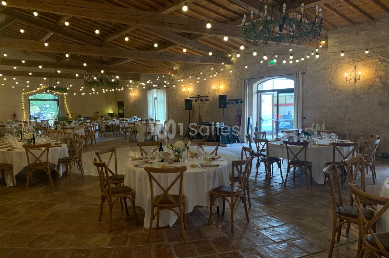 Salle de réception décorée avec des guirlandes lumineuses, tables rondes dressées et chaises en bois.