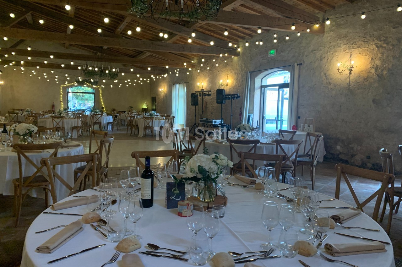 Salle de réception décorée avec des guirlandes lumineuses, tables dressées avec nappes blanches, fleurs et couverts.