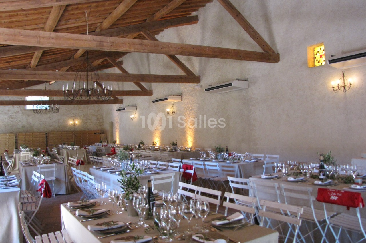 Salle de réception avec tables dressées, chaises blanches, poutres apparentes et éclairage chaleureux.