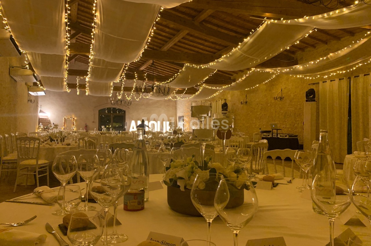 Salle de réception décorée avec des guirlandes lumineuses, tables dressées avec vaisselle et centre de table floral.