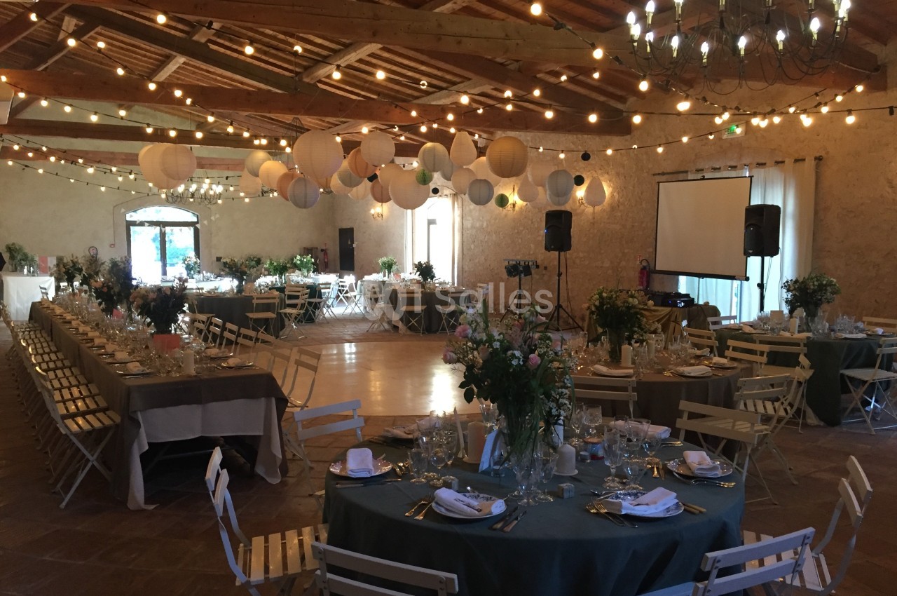 Salle de réception décorée avec des guirlandes lumineuses, des lanternes suspendues et des tables dressées pour un événement.