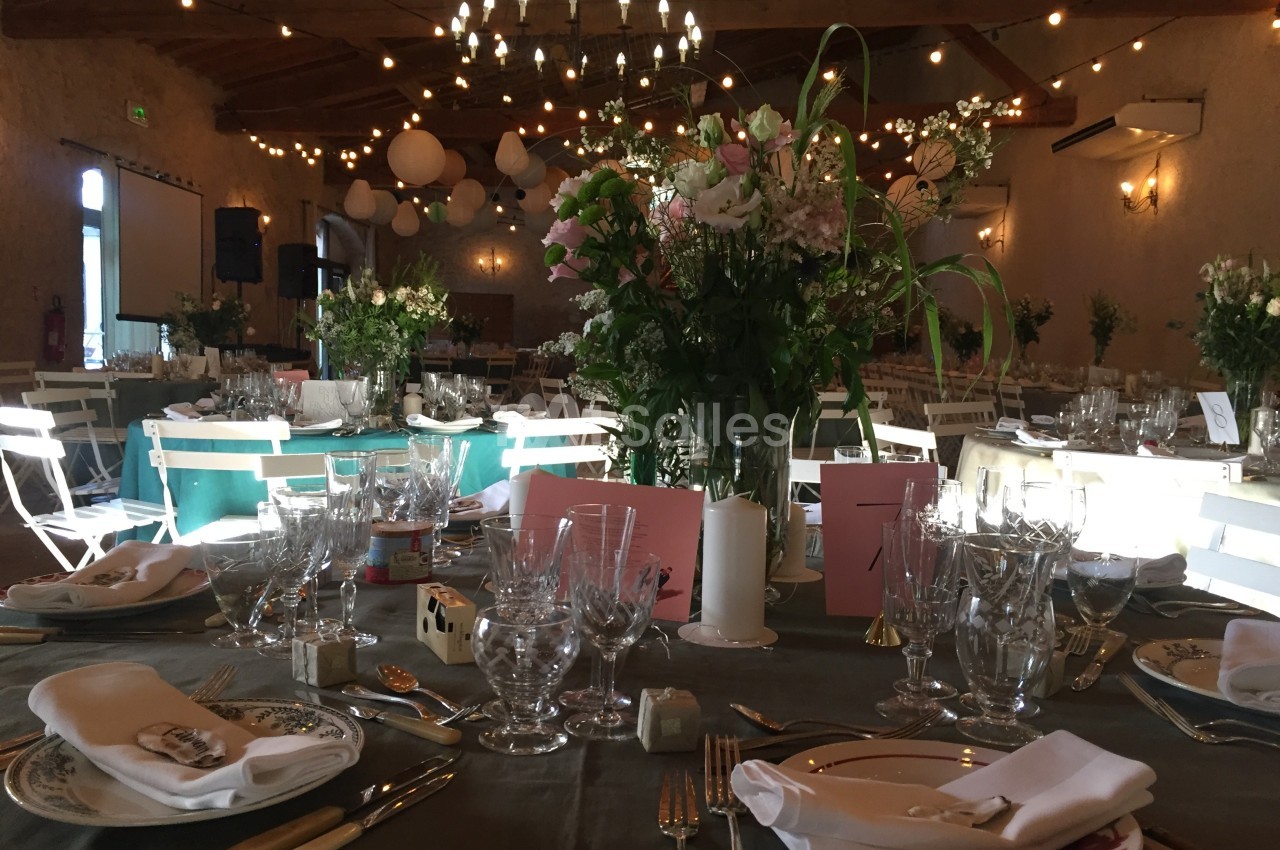 Salle de réception décorée pour un événement, avec tables dressées, fleurs, vaisselle et guirlandes lumineuses suspendues.