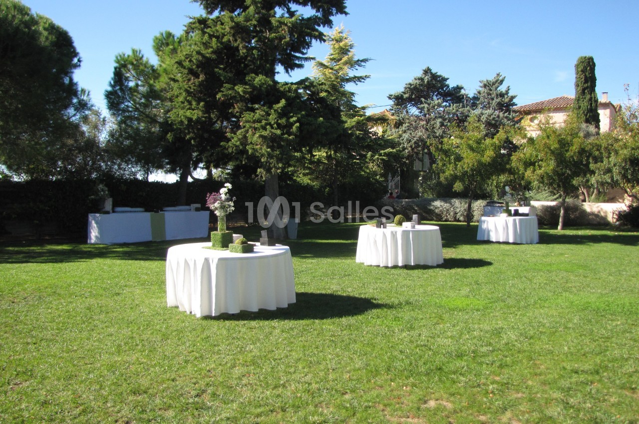 Tables rondes dressées avec nappes blanches sur une pelouse, entourées d'arbres et de végétation sous un ciel dégagé.