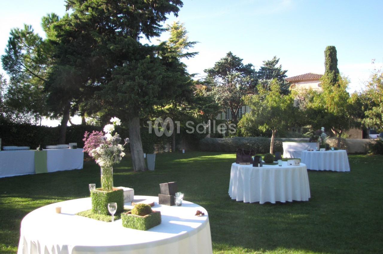 Tables rondes dressées avec nappes blanches dans un jardin verdoyant, entouré d'arbres et de végétation.