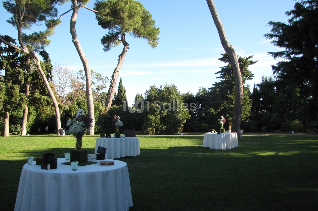 Tables rondes dressées avec nappes blanches sur une pelouse, entourées d'arbres sous un ciel dégagé.
