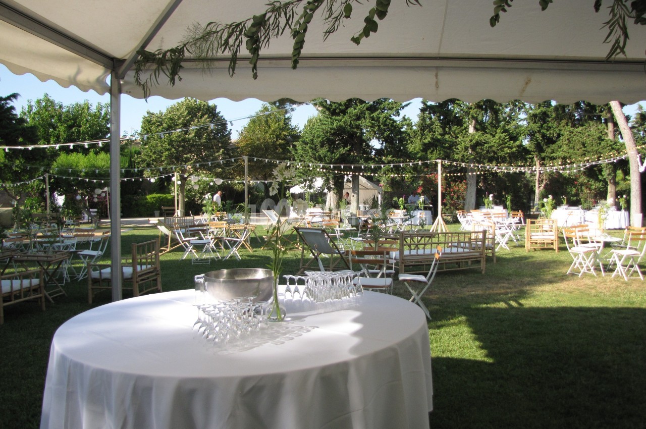 Tables dressées sous une tente dans un jardin, avec guirlandes lumineuses suspendues et végétation en arrière-plan.