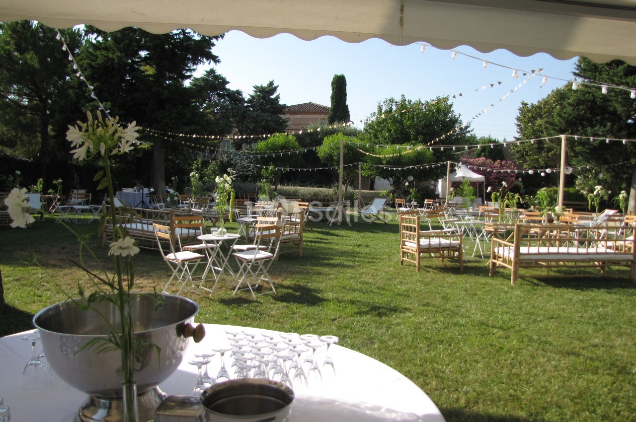 Espace extérieur aménagé pour un événement avec tables, chaises, guirlandes lumineuses et verdure environnante.