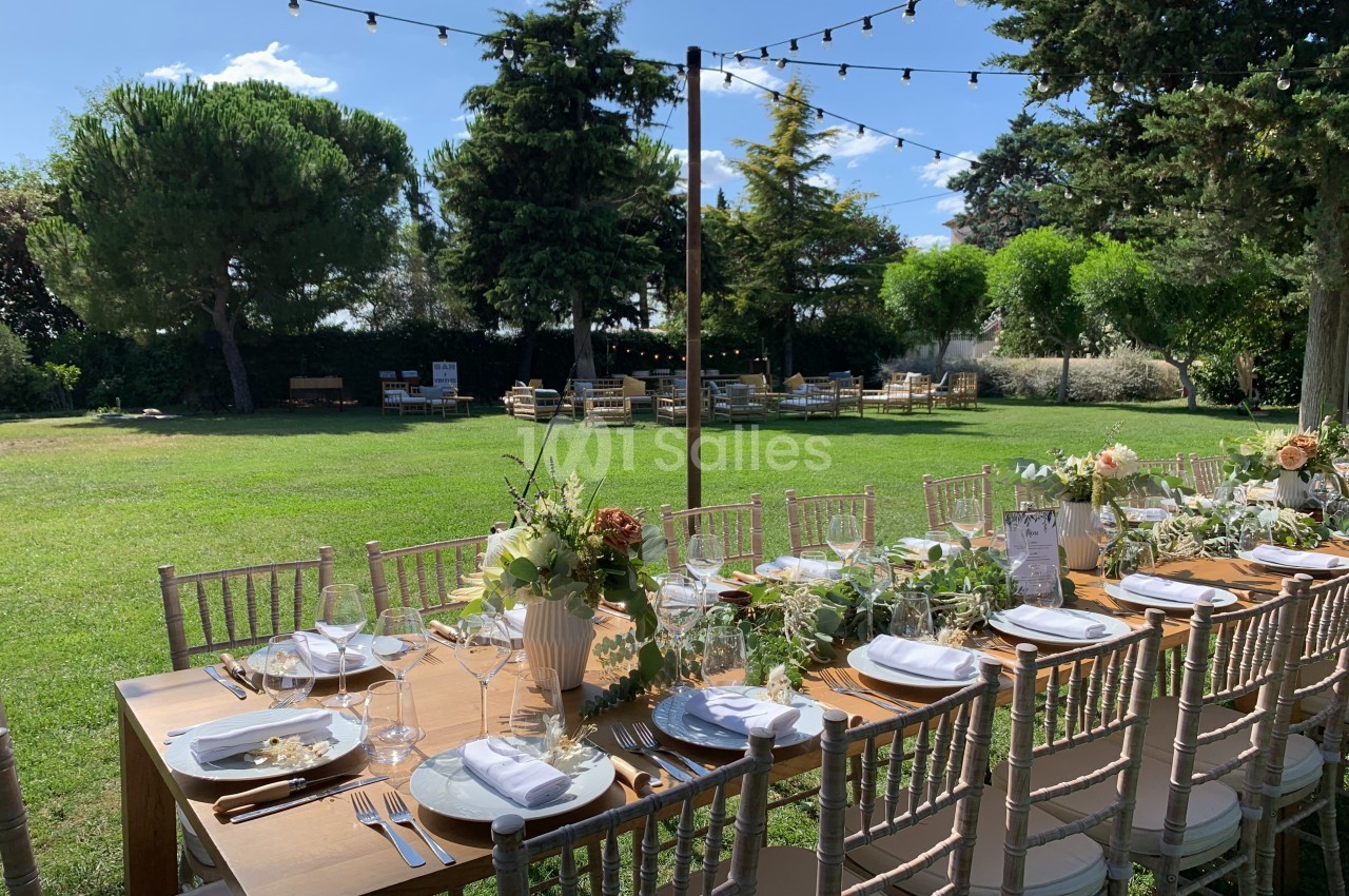 Table en bois décorée pour un repas en plein air, entourée de chaises, dans un jardin avec des arbres et guirlandes…