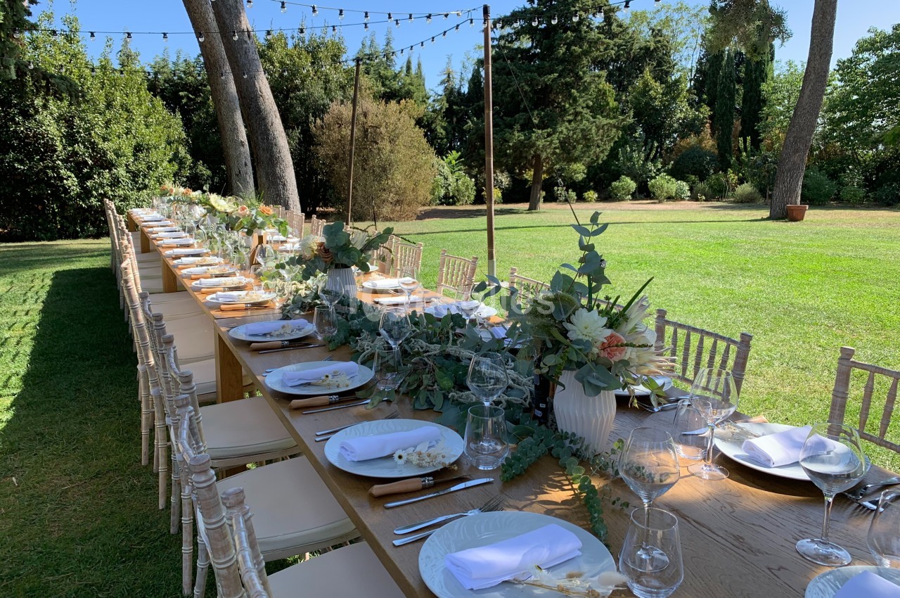 Grande table en bois décorée de fleurs et vaisselle élégante, installée dans un jardin sous des guirlandes lumineuses.