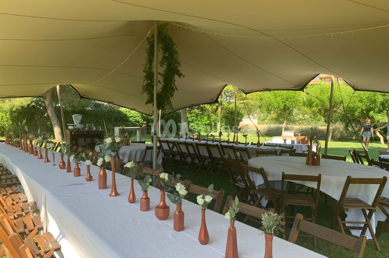 Tables longues décorées de vases en cuivre et fleurs, disposées sous une tente blanche dans un jardin verdoyant.