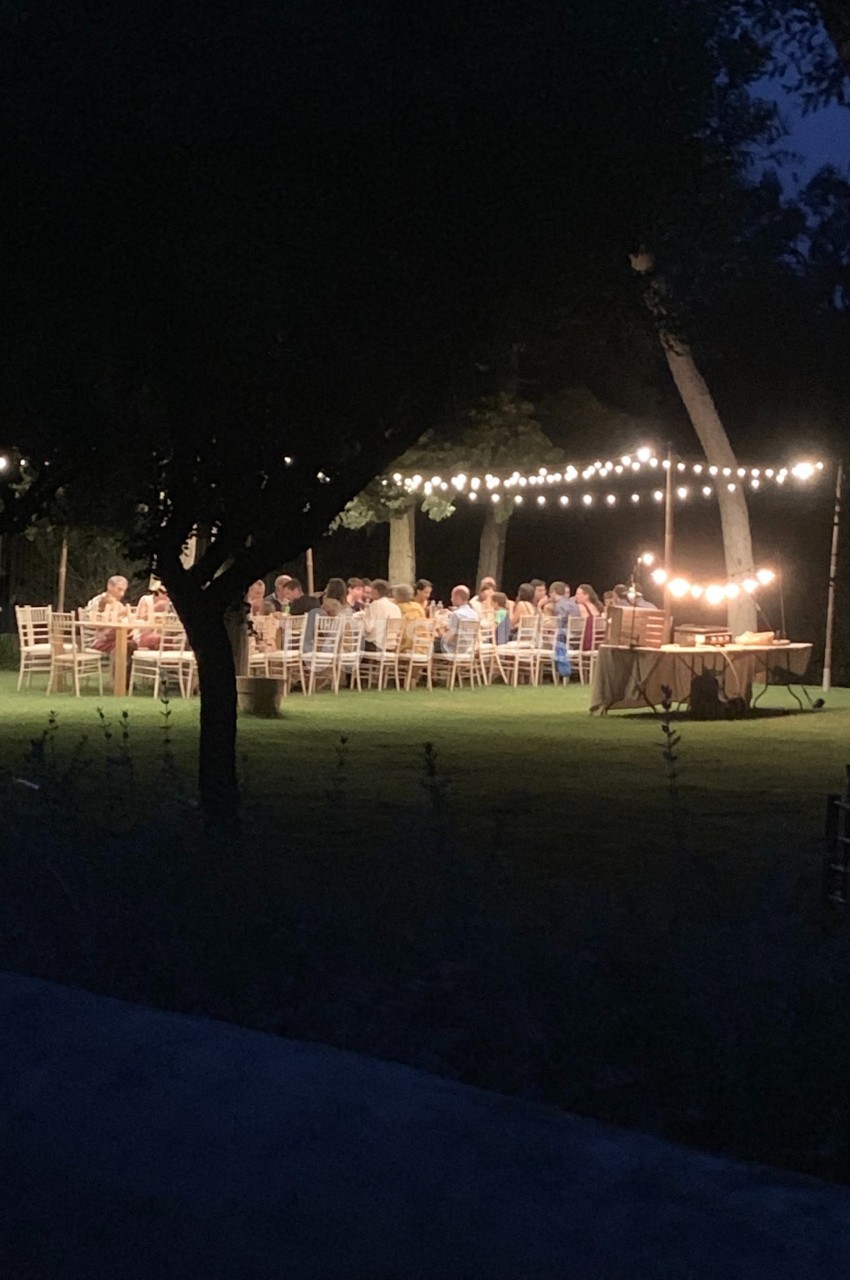 Des personnes dînent en plein air sous des guirlandes lumineuses dans un jardin, en soirée.