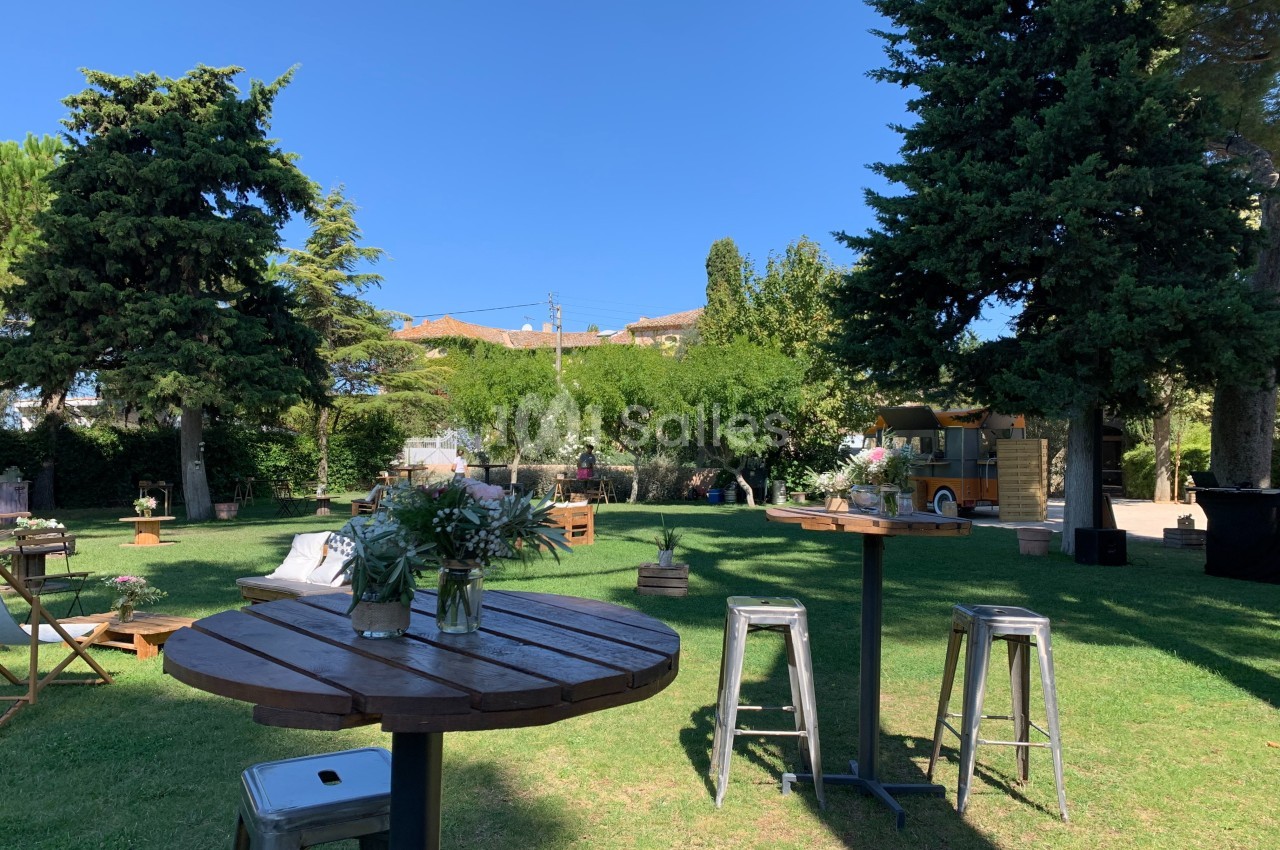 Espace extérieur avec tables hautes, tabourets et décorations florales, entouré d'arbres et de verdure sous un ciel bleu.