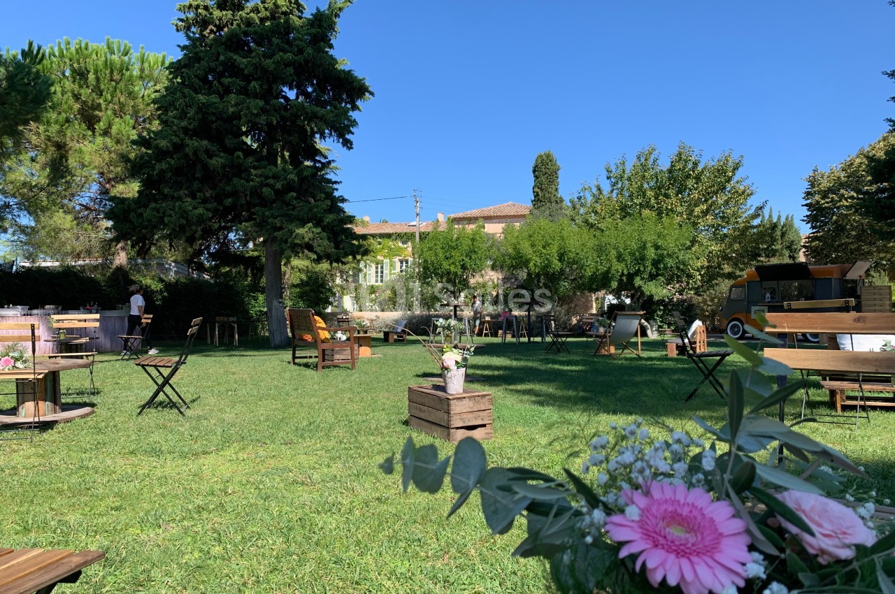 Espace extérieur avec tables et chaises sur une pelouse, décoré de fleurs, entouré d'arbres et de bâtiments.