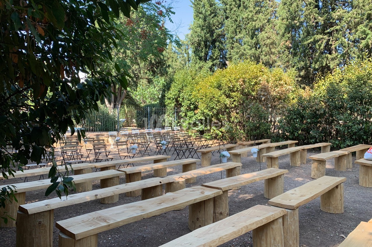 Bancs en bois disposés en rangées dans un espace extérieur entouré de végétation et de chaises pliantes.