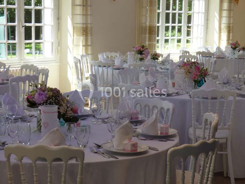 Salle lumineuse avec tables rondes dressées pour un événement, décorées de fleurs et de serviettes pliées.