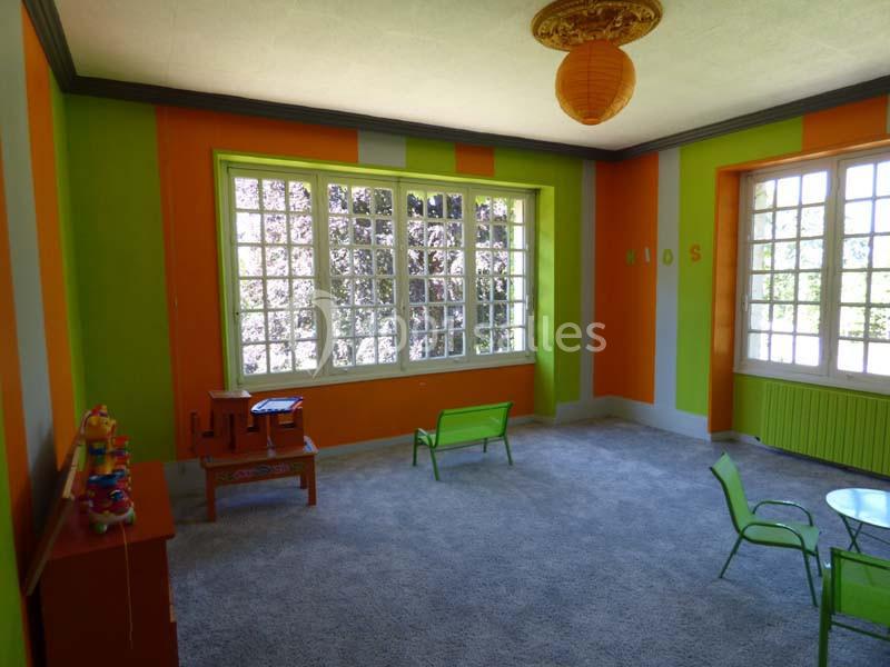 Salle de jeux lumineuse avec murs colorés orange et vert, mobilier pour enfants et grandes fenêtres donnant sur l'extérieur.