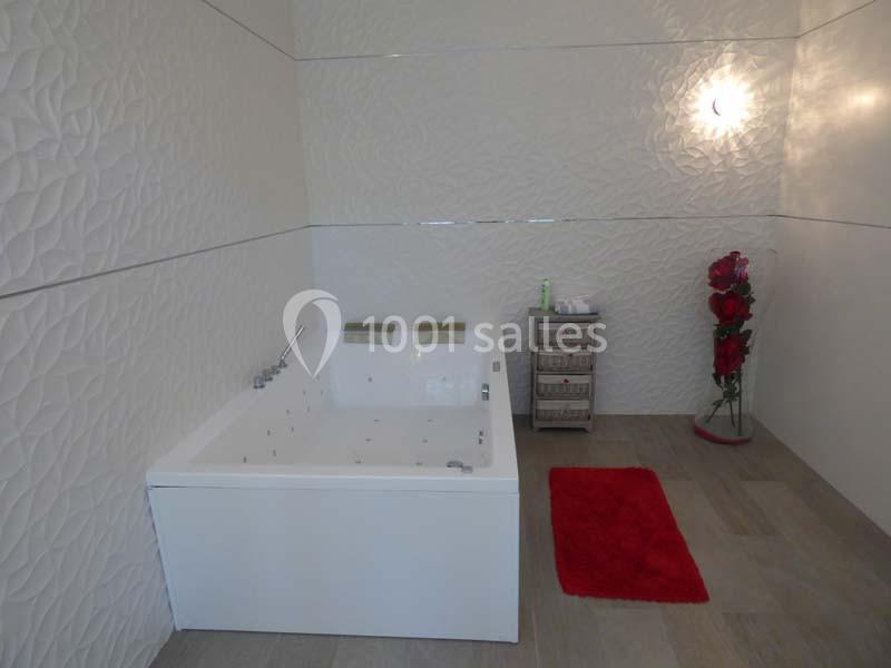 Salle de bain moderne avec une baignoire blanche, un tapis rouge, des murs texturés et une décoration florale.