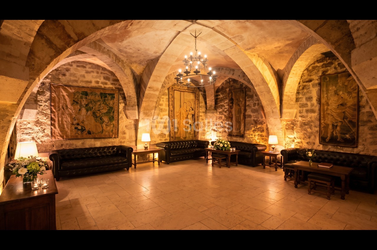 Salle voûtée en pierre avec canapés en cuir, tapisseries murales, tables en bois et éclairage chaleureux.