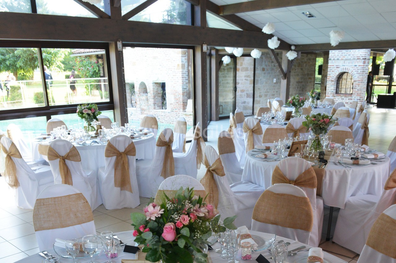 Salle de réception décorée avec des tables rondes, nappes blanches, chaises ornées de nœuds en toile de jute et fleurs.