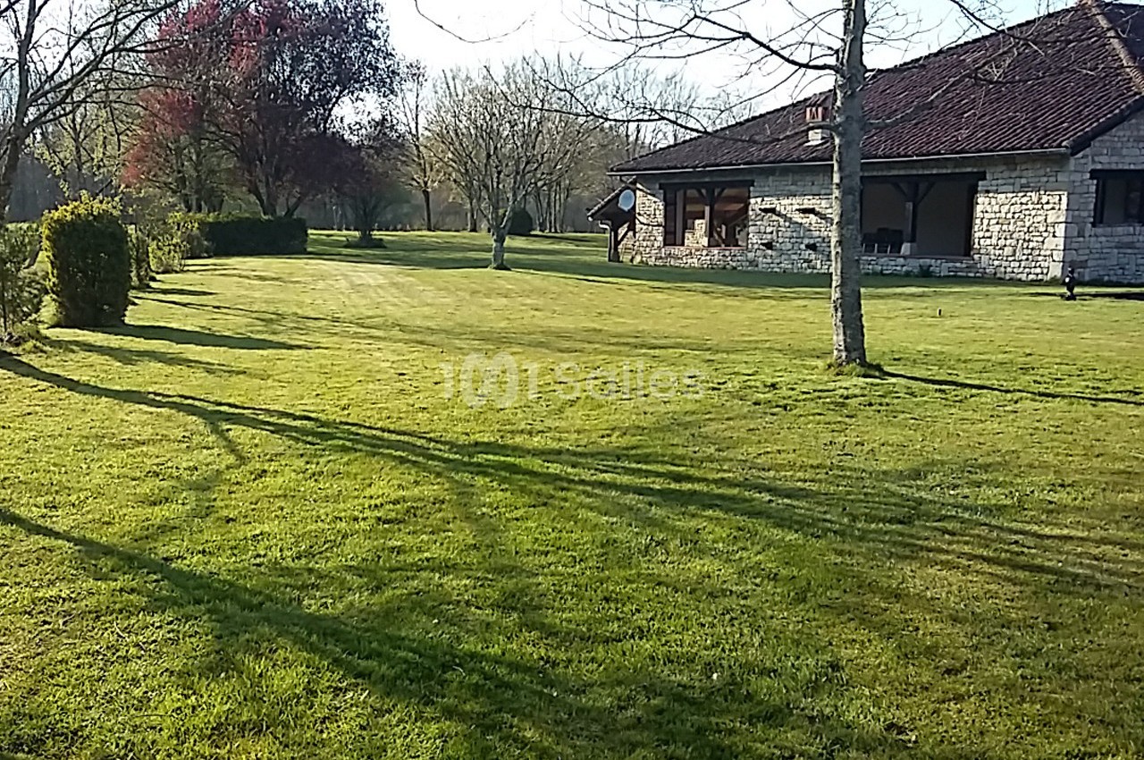 Pelouse verdoyante avec arbres et une maison en pierre au toit brun sous un ciel dégagé.