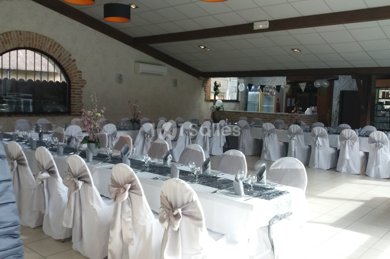 Salle de réception décorée pour un événement, avec des tables dressées et des chaises recouvertes de housses blanches.