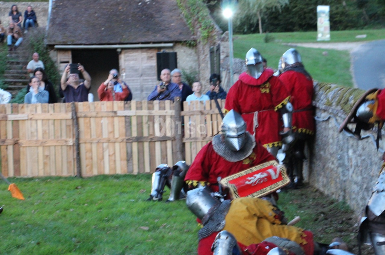 Des personnes en armures médiévales participent à une reconstitution historique devant un public près d'une palissade.